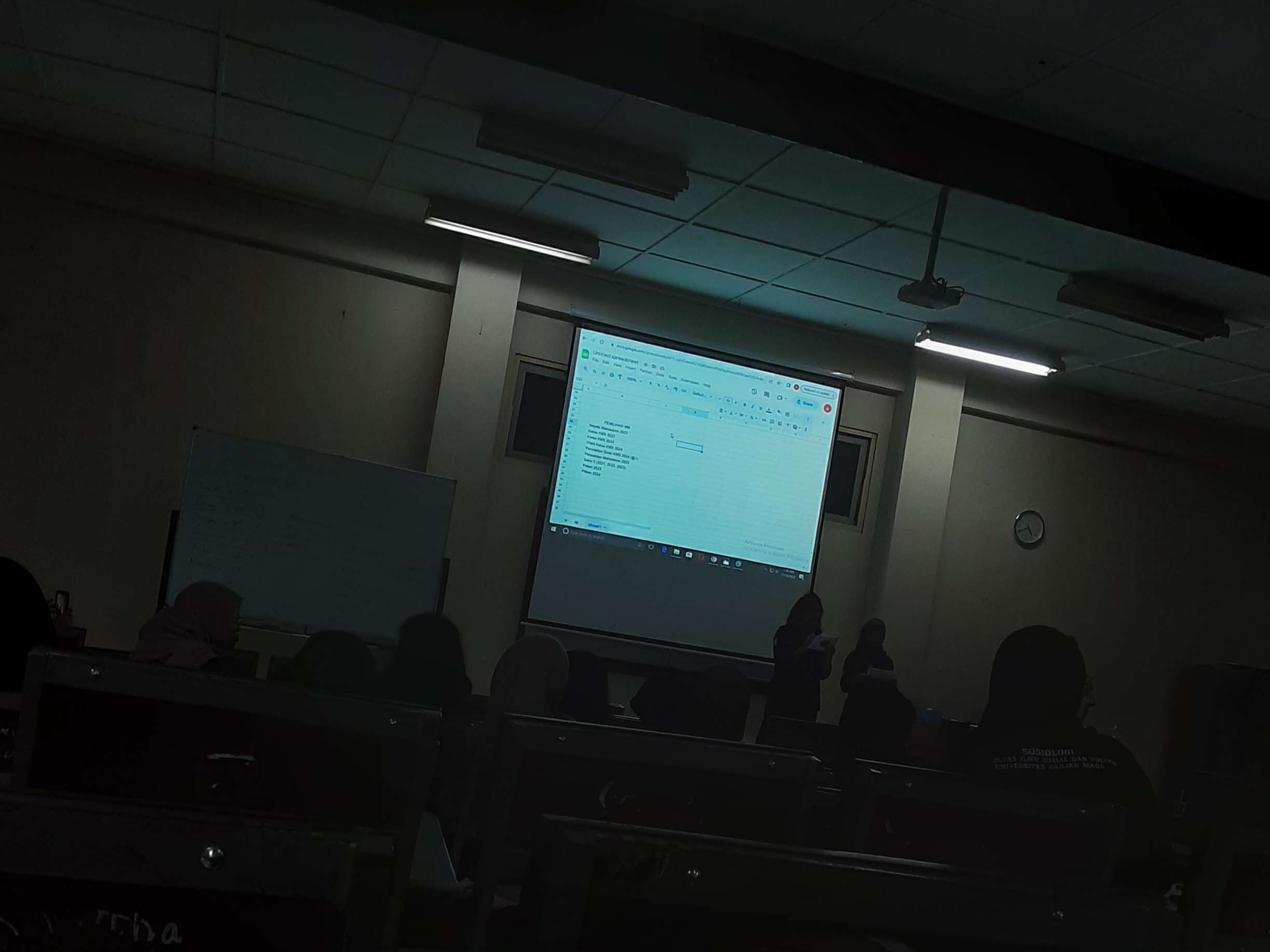 Foto dengan situasi gelap, fokus dengan presentasi yang ditampilkan melalui proyektor.